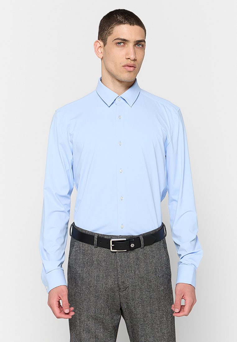 Chemise à manches longues bleu clair avec un col pointu et des boutons à l'avant, portée avec un pantalon à motifs gris et une ceinture noire.