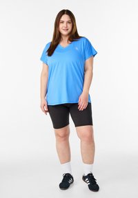 Hellblaues Performance-Oberteil mit kurzen Ärmeln und V-Ausschnitt. Kombiniert mit schwarzen Fahrradshorts. Das Model trägt dunkle Turnschuhe und weiße Socken.