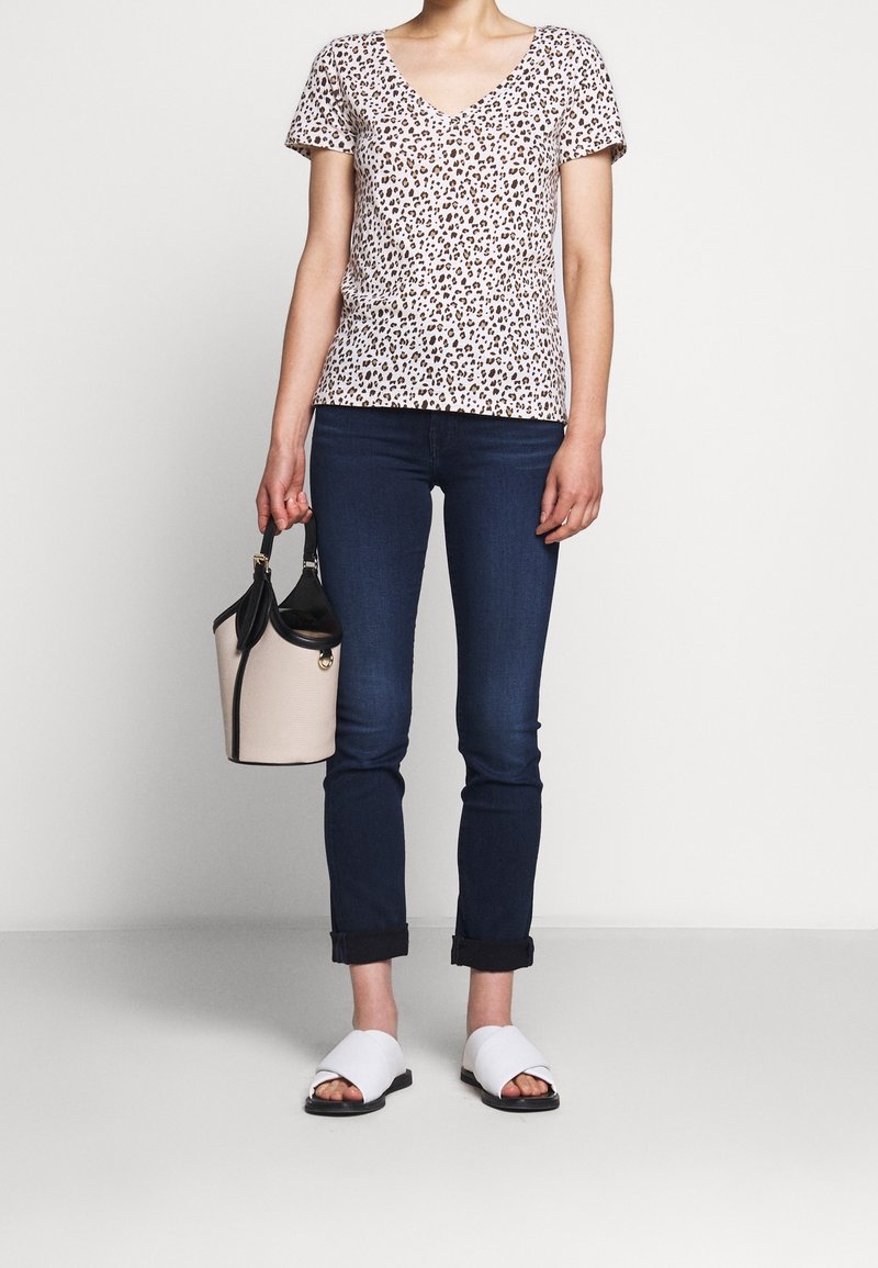 Femme portant une chemise à manches courtes imprimé léopard, un jean foncé retroussé, des sandales blanches à enfiler, tenant un sac à main beige et noir.