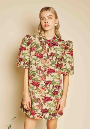 Jeune femme portant une robe à motif floral avec des manches bouffantes et un col à nœud, posant devant un fond beige.