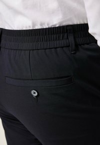 Pantaloni neri con girovita elastico, dotati di una tasca posteriore chiusa da un bottone. Il tessuto ha una texture liscia e un design sartoriale.