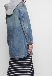 Veste en denim avec un délavage bleu clair, à manches longues et à coupe décontractée. Fini avec une capuche grise, des coutures visibles et une légère usure.