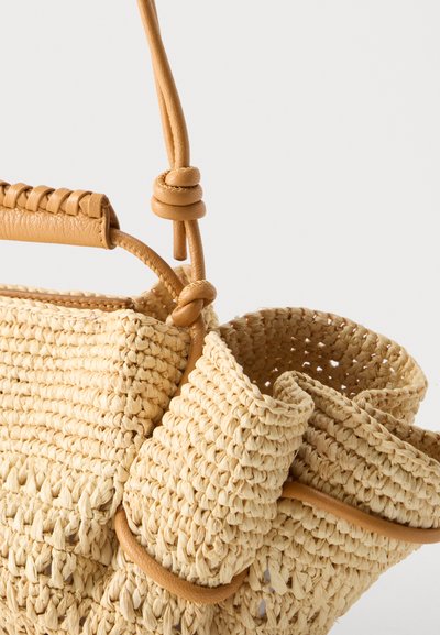 Κοντινή λήψη μιας μπεζ υφαντής ψάθινης τσάντας με δερμάτινο λουρί και δεμένες δερμάτινες λεπτομέρειες σε απλό λευκό φόντο.
