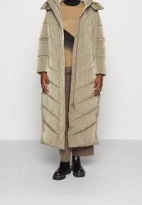 Abrigo de plumas beige con capucha y diseño de acolchado en forma de chevron. Presenta un cierre frontal de cremallera y cierres de botón. Llevado sobre una blusa marrón y pantalones.