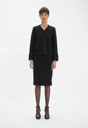 Costume noir en deux pièces comprenant une veste cintrée avec des finitions texturées et une jupe longueur genou. Fabriqué en tissu lisse, des bottes complètent le look.