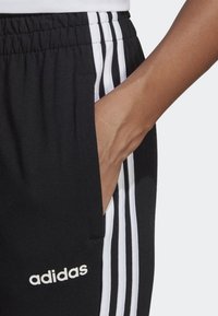 Svarta träningbyxor med tre vita ränder längs sidan, elastisk midjeband och en sidoficka. Har en vit "adidas"-logotyp.