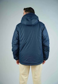 Doudoune bleu marine avec capuche, présentant une texture lisse, des poignets élastiques et une coupe légèrement oversize. Vue arrière avec des détails minimalistes.