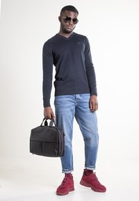 Maglione nero a scollo a V, jeans di denim azzurro chiaro e scarpe da ginnastica rosse. Tiene una borsa in pelle nera con due manici superiori e chiusura con zip.