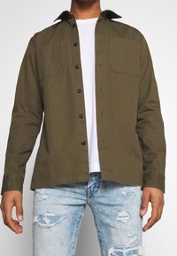 Camisa verde oliva con botones, cuello puntiagudo, dos bolsillos en el pecho y botones negros, llevada sobre una camiseta blanca y unos vaqueros claros de mezclilla.