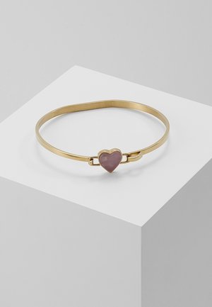 Gouden armband met een roze hartvormige steen als middelpunt, rustend op een wit geometrisch oppervlak.