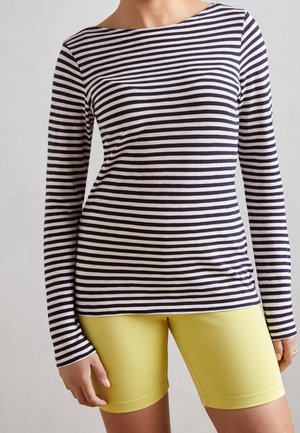 Persoon in een zwart-wit gestreept shirt met lange mouwen en felgele, aansluitende shorts, staand tegen een effen achtergrond.