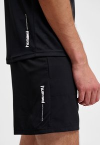 Svart träningsskjorta och shorts i lättviktsmaterial, med vita "hummel performance"-logotyper på sidorna. Närbild.