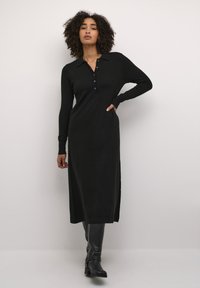 Black long-sleeve dress with a collar, featuring button detailing down the front and a straight cut, worn with knee-high black boots.
