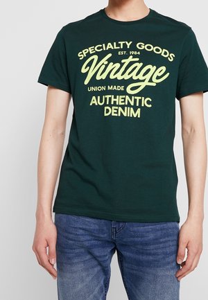Osoba nosząca ciemnozieloną koszulkę z żółtym napisem "Specialty Goods Vintage Union Made Authentic Denim" i niebieskie dżinsy.