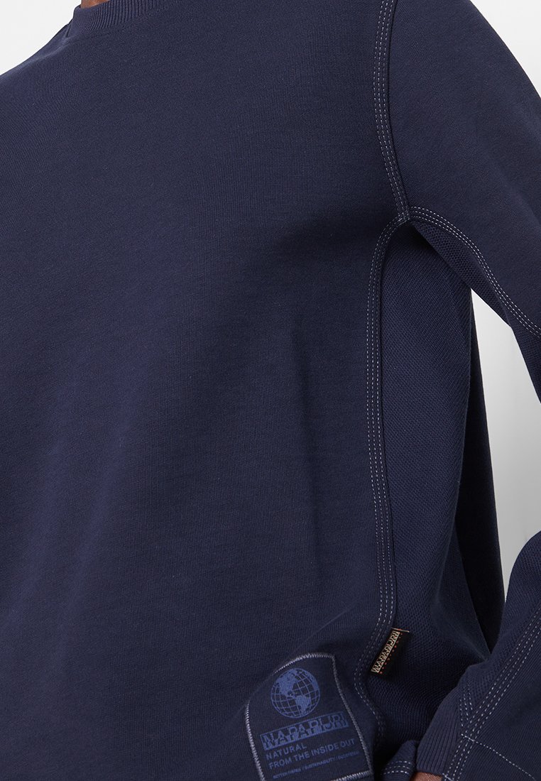 Marineblaue Langarmshirt mit sichtbaren Doppelstichen und einem kleinen rechteckigen Aufnäher nahe dem Saum, der einen Globus und den Markennamen zeigt.