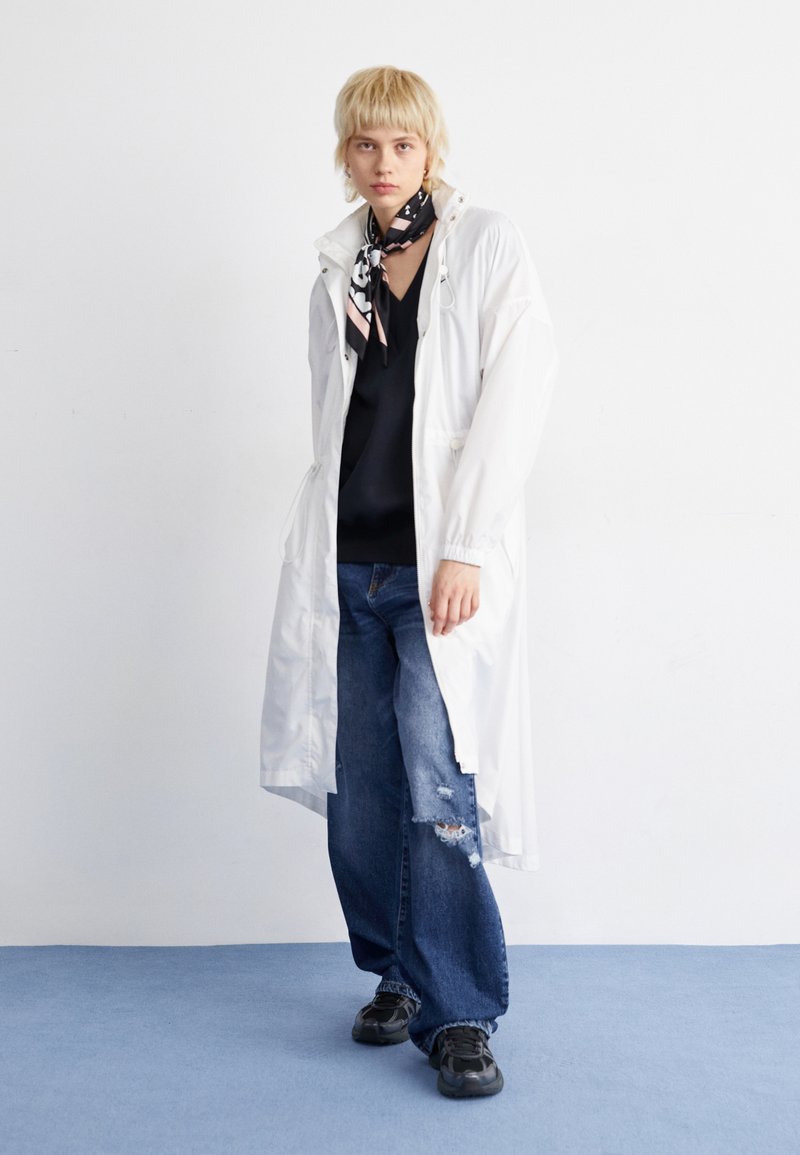 Lange witte regenjas van lichtgewicht materiaal, met een trekkoord in de taille en een capuchon, gecombineerd met donkere jeans en zwarte sneakers.