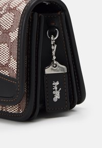 Brązowa torebka wykonana z plecionego materiału z czarnymi skórzanymi akcentami, z metalowymi elementami w kolorze srebrnym i zawieszką. Szyte detale podkreślają design.