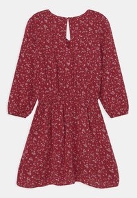 Vestido floral rojo con mangas largas, cintura fruncida y un detalle de abertura en la parte de atrás. Presenta pequeños patrones de flores blancas y lavanda.