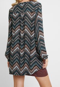 Blouse à manches longues de couleur bleu sarcelle foncé avec un motif en zigzag en marron, crème et bleu clair. Coupe décontractée avec un détail froncé au dos.