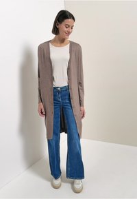 Dlouhý hnědý kardigan z měkké látky, s knoflíky vpředu, nošený přes bílý top a modré zvonové džíny, doplněný bílými teniskami.