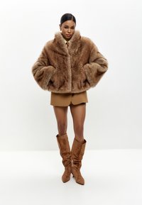 Brun faux fur cropped jakke med brede ærmer, der har en blød tekstur og en stor krave, parret med et brunt shorts sæt og matchende støvler.