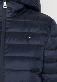 Giacca trapuntata blu navy con cuciture orizzontali, zip frontale e piccolo logo rettangolare rosso, bianco e blu sul lato sinistro del petto.