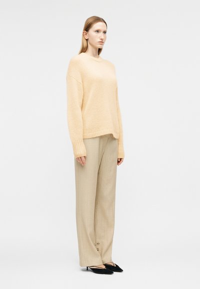 Femme debout portant un pull oversize beige doux, un pantalon ample beige clair avec des détails à boutons, et des talons noirs pointus.