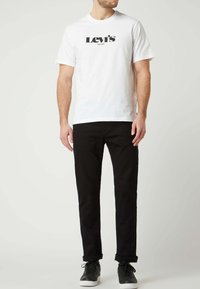 Camiseta blanca de algodón con el logo negro de "Levi's", combinada con pantalones vaqueros negros. La modelo lleva zapatillas negras y está de pie sobre un fondo neutro.