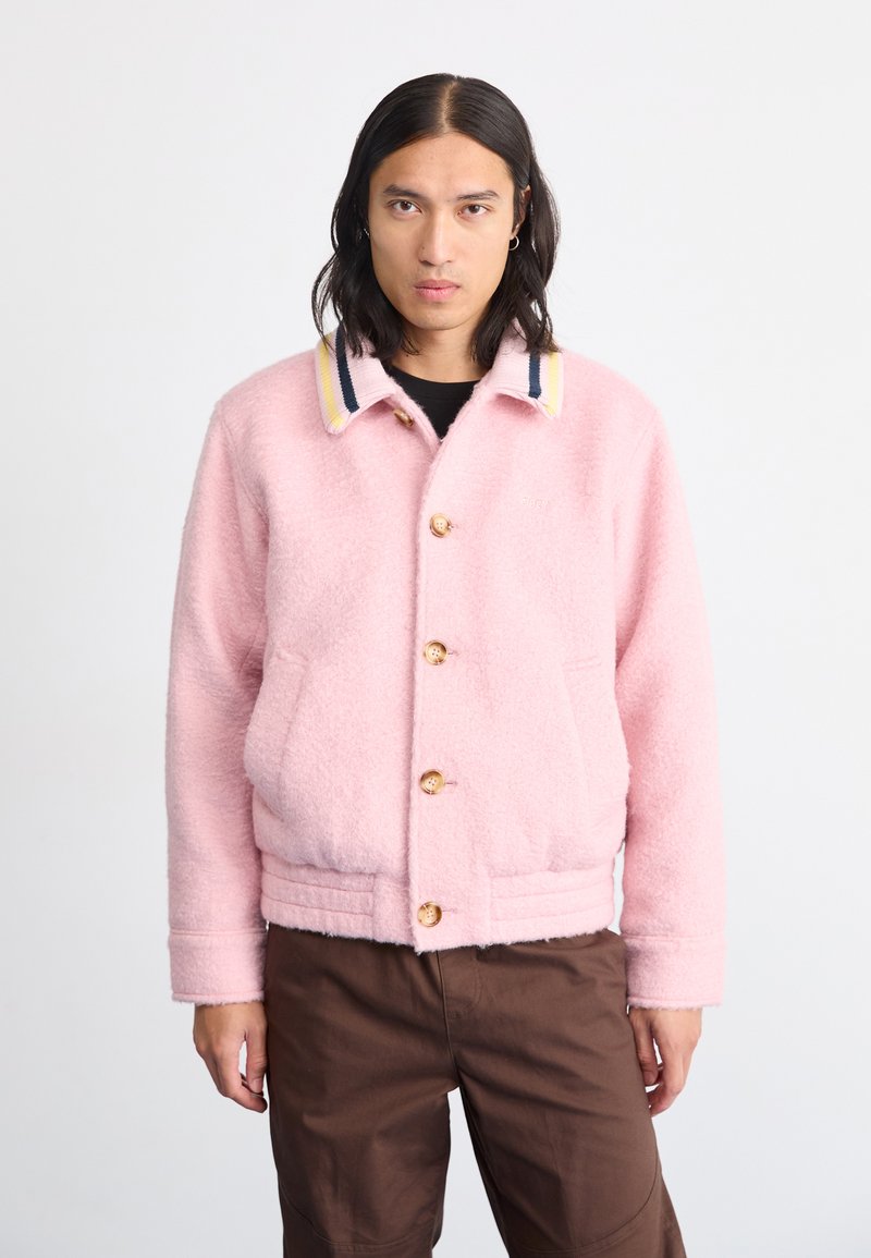 Casaco de fleece rosa com colarinho, apresentando riscas amarelas, azuis marinhas e brancas, fecho de botão de madeira e cintura elástica; usado com calças castanhas.