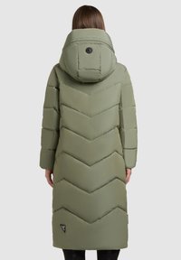 Ilgas alyvuogių žalios pūkų kailis su chevrono siuvinėjimo raštu, aukštu apykakle, didele kapuce ir matoma prekės ženklu šalia krašto.