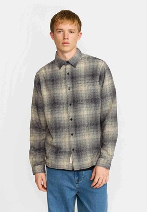 Jeune homme portant une chemise beige et noire à carreaux avec boutons et un jean bleu, debout devant un fond clair uni.