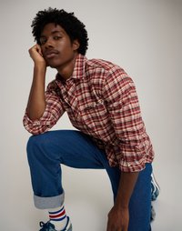 Červená a krémová kockovaná košeľa s gombíkmi, modré džínsy s ohrnutými rukávmi a viacfarebné pruhované ponožky. Neformálny postoj, neutrálny pozadie.