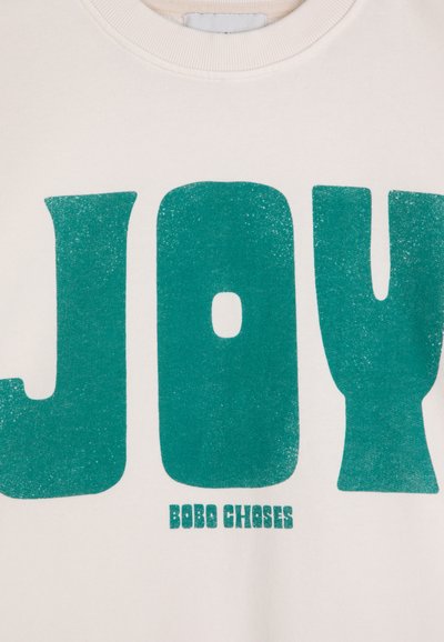 Felpa color crema con grandi lettere teal che scrivono "JOY" e il nome del marchio "BOBO CHOSES" in un testo teal più piccolo sottostante.