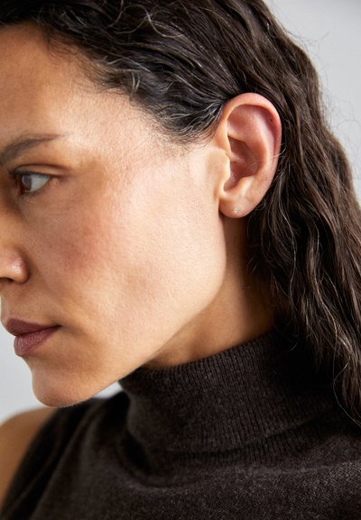 Primer plano de el perfil de una mujer con cabello oscuro y ondulado, piel marrón clara y un suéter de cuello alto marrón. Unos simples pendientes de botón acentúan su oreja.