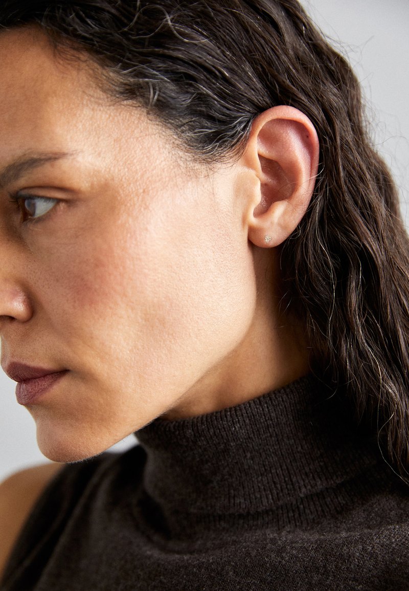 Zbliżenie profilu kobiety z ciemnymi, falującymi włosami, jasnobrązową skórą i brązowym golfem. Prosta kolczyk-wkrętka podkreśla jej ucho.