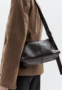 Borsa a tracolla nera in pelle con una forma elegante, caratterizzata da dettagli di cucitura minimali e una tracolla regolabile, indossata sopra un giubbotto marrone.