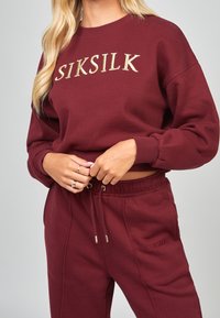 Sudadera de algodón burdeos con el logotipo "SIKSILK" bordado en dorado, escote redondo y puños acanalados, combinada con pantalones a juego con cordón.