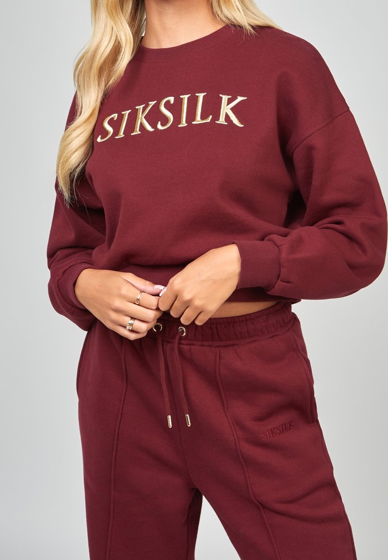 Sudadera de algodón burdeos con el logotipo "SIKSILK" bordado en dorado, escote redondo y puños acanalados, combinada con pantalones a juego con cordón.