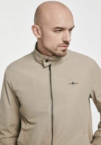 Beżowa, lekka kurtka z wysokim kołnierzem, przednim zamkiem błyskawicznym oraz dyskretnym logo na lewej piersi, charakteryzująca się gładką fakturą.