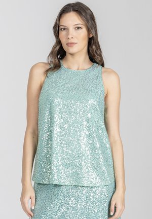 Kobieta ubrana w bezrękawnik z błyszczących, jasnoniebieskich cekinów oraz pasującą spódnicę, stojąca na tle jednolitego szarego tła.