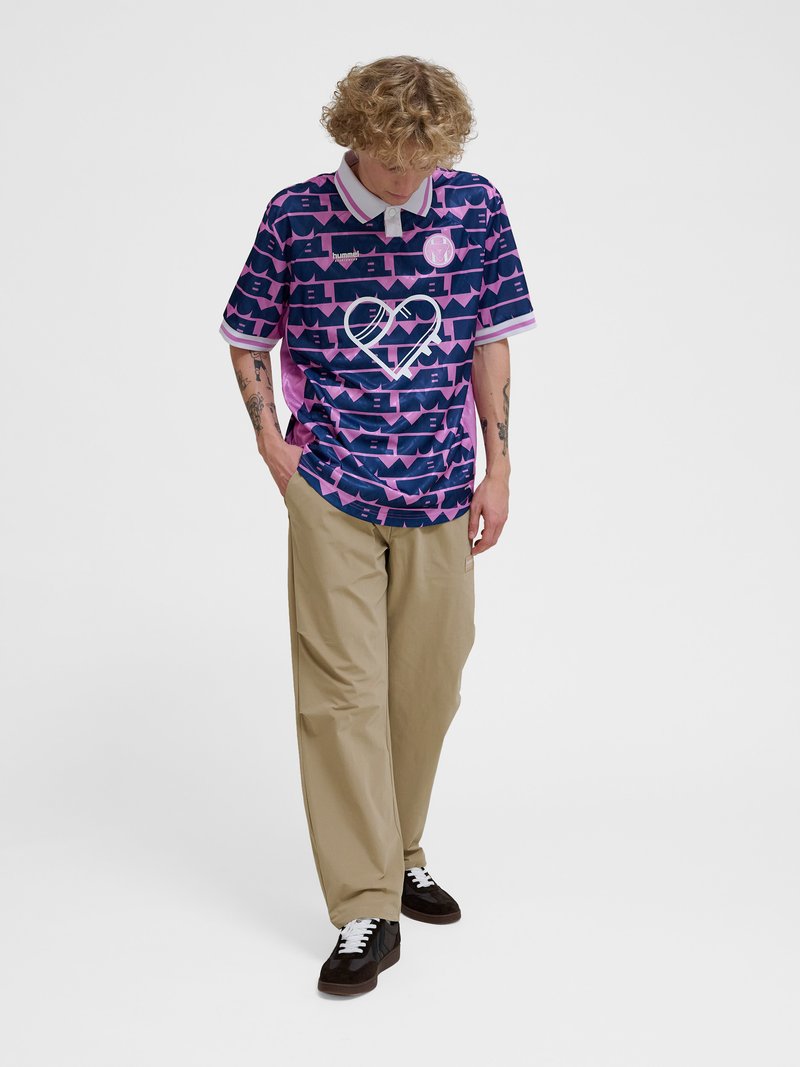 Młoda osoba z kręconymi włosami nosi fioletową, wzorzystą koszulę z krótkim rękawem, beżowe spodnie i czarne trampki, patrzy w dół z rękami w kieszeniach.