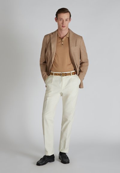 Blazer beige sur un polo marron, pantalon blanc et chaussures noires. La ceinture présente un design tressé en marron et beige. Texture lisse dans l'ensemble.