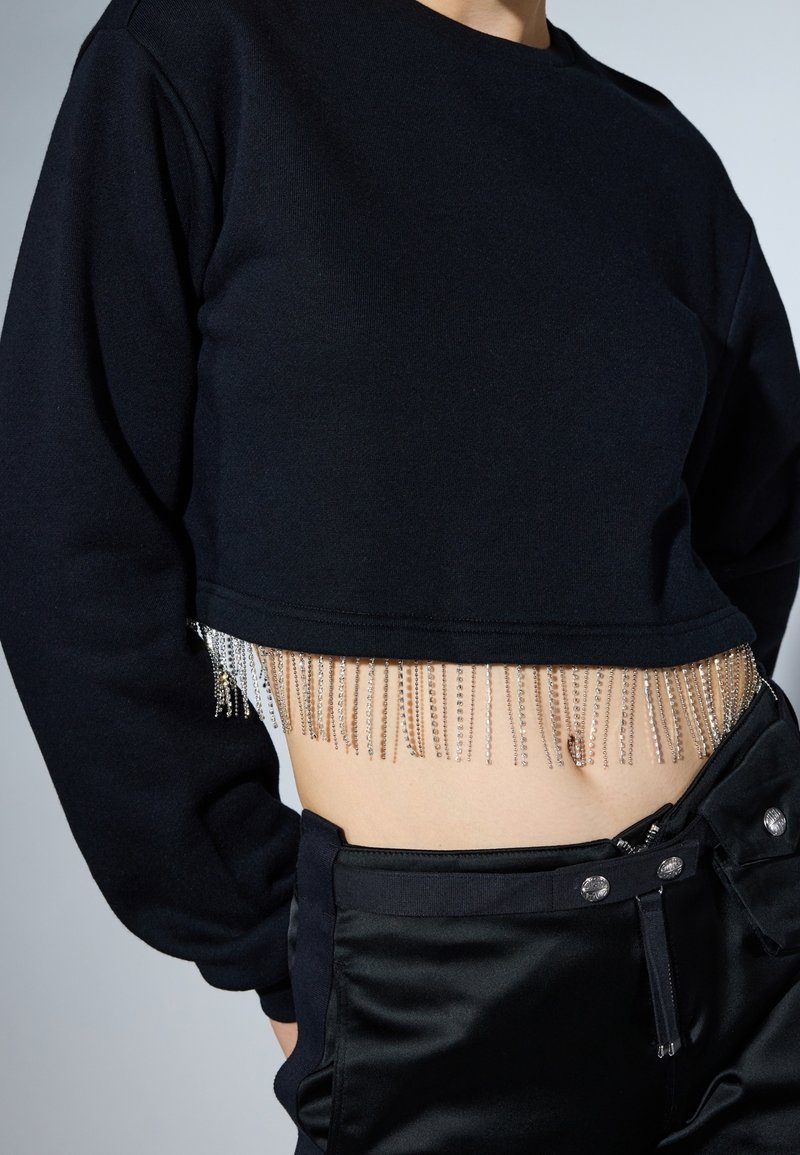 Personne portant un sweat court noir avec des franges argentées à l’ourlet et un pantalon noir taille haute avec une poche boutonnée.