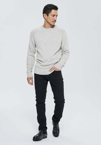 Homme marchant portant un pull gris clair moucheté, un pantalon noir et des chaussures en cuir noires sur un fond clair.