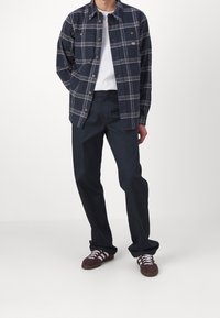 Camicia di flanella a quadri blu navy con bottoni, due tasche sul petto, t-shirt bianca sotto, pantaloni scuri e sneakers bordeaux con strisce bianche.