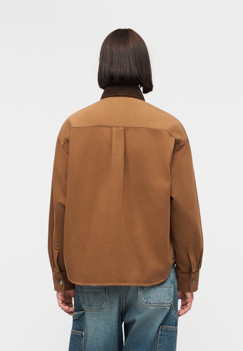 Persona con capelli lisci e scuri che indossa una giacca marrone e jeans blu, in piedi di spalle su uno sfondo chiaro e uniforme.