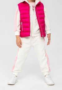 Gilet in piuma rosa sopra un felpa bianca e pantaloni della tuta con strisce rosa ai lati. Sneakers bianche completano l'outfit, mettendo in risalto i strati e il contrasto di colori.