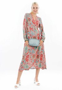Donna che indossa un vestito a wrap floreale colorato con maniche lunghe, sandali con tacco bianchi e una borsa a tracolla azzurro chiaro, in piedi su uno sfondo bianco.