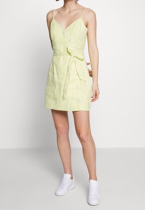 Frau trägt ein ärmelloses, hellgelbes Minikleid mit Knöpfen und seitlicher Schleife, kombiniert mit weißen Sneakers vor einfarbigem Hintergrund.