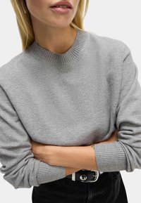 Szary sweter z prążkowanym okrągłym dekoltem i prążkowanymi mankietami. Miękka faktura, casualowy design, w połączeniu z czarnym paskiem z metalową klamrą.